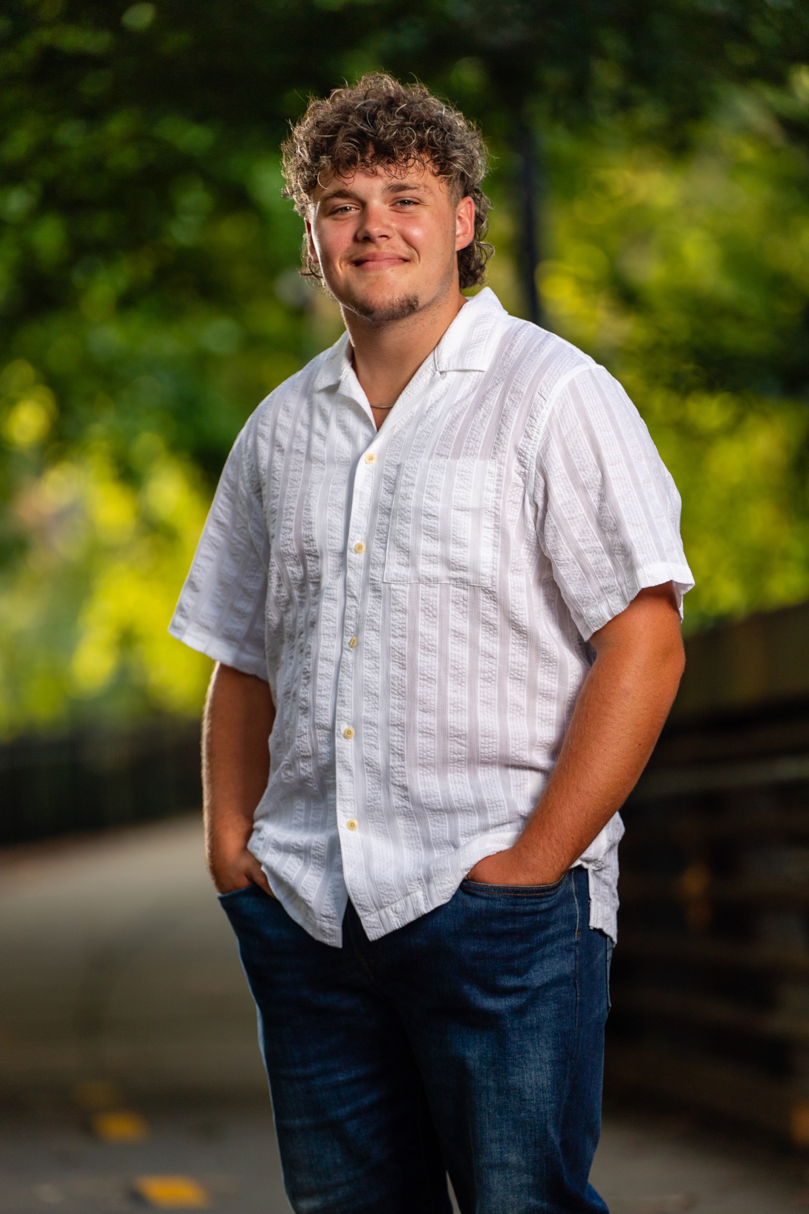A high school senior boy in a white button-up shirt and jeans standing with his hands in his pockets on a tree-lined path in golden evening light.