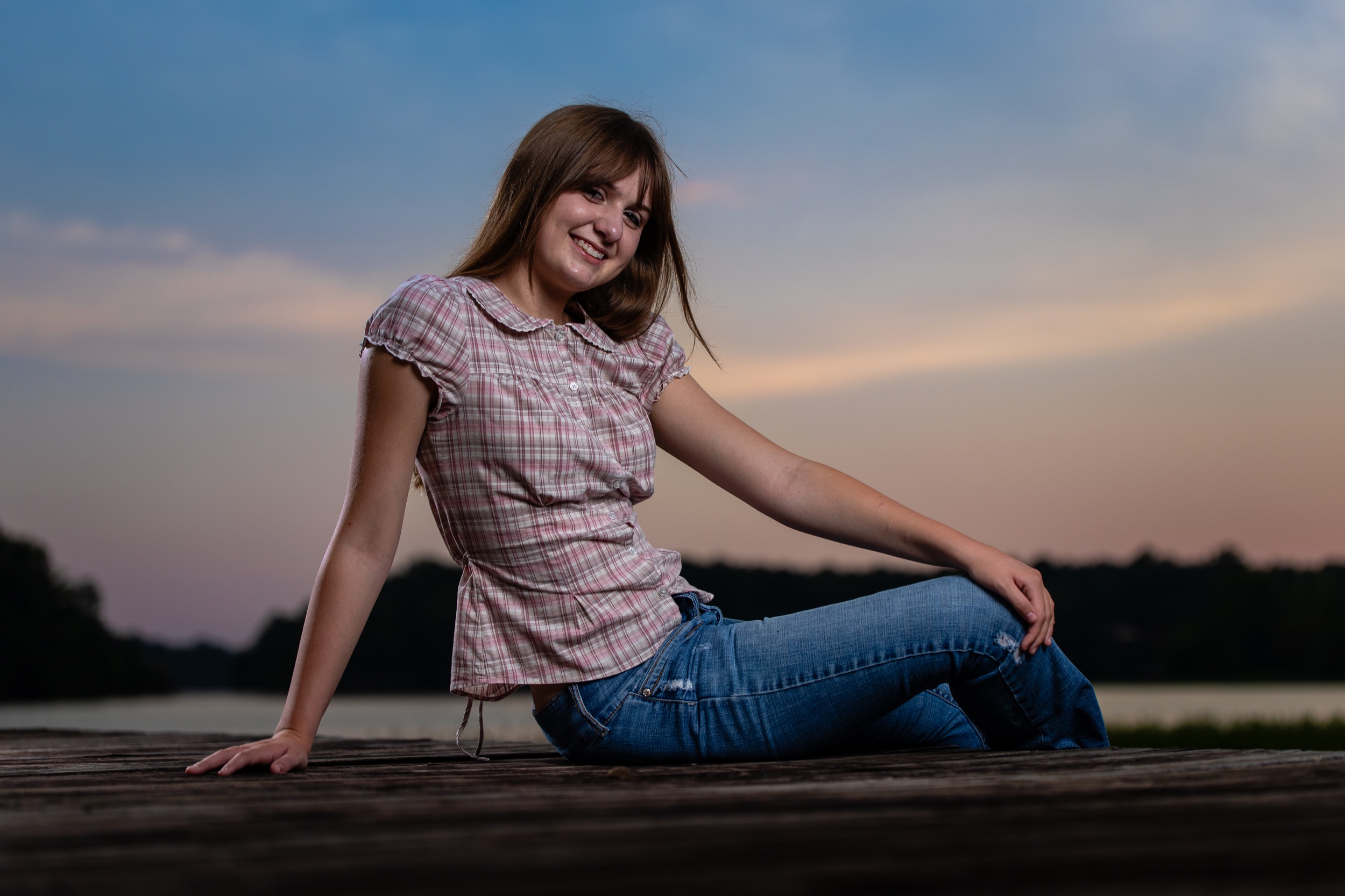 A high school senior girl in a pink plaid shirt and jeans sitting on a wooden dock at dusk, a soft sunset over the tree line behind her.